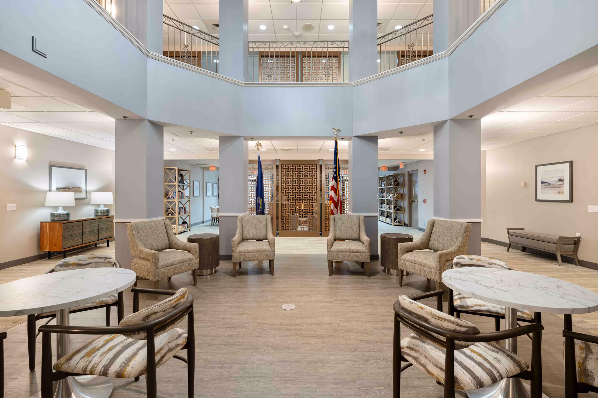 A spacious and well-lit common area in a senior living community featuring four beige armchairs arranged in a semi-circle around two small round tables. In the foreground, there are two marble-top tables each with two striped cushioned chairs. The room has light-colored walls, wooden flooring, and decorative shelving units along the walls. An American flag and another flag stand near a decorative wooden panel at the back. The area has a high ceiling with a balcony railing visible above.