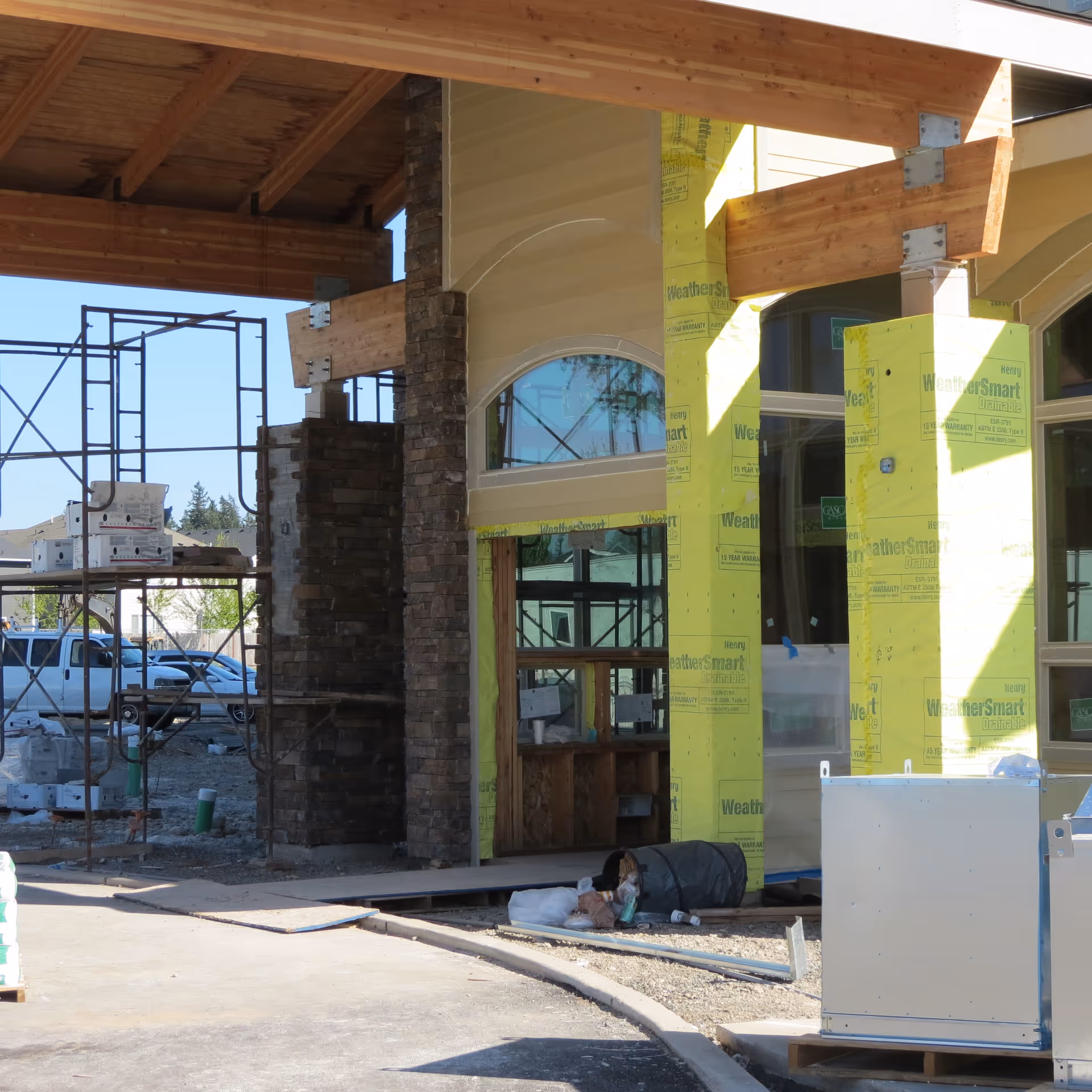 Front entrance of a senior living building under construction with exposed yellow sheathing, stone columns, scaffolding, and construction materials.