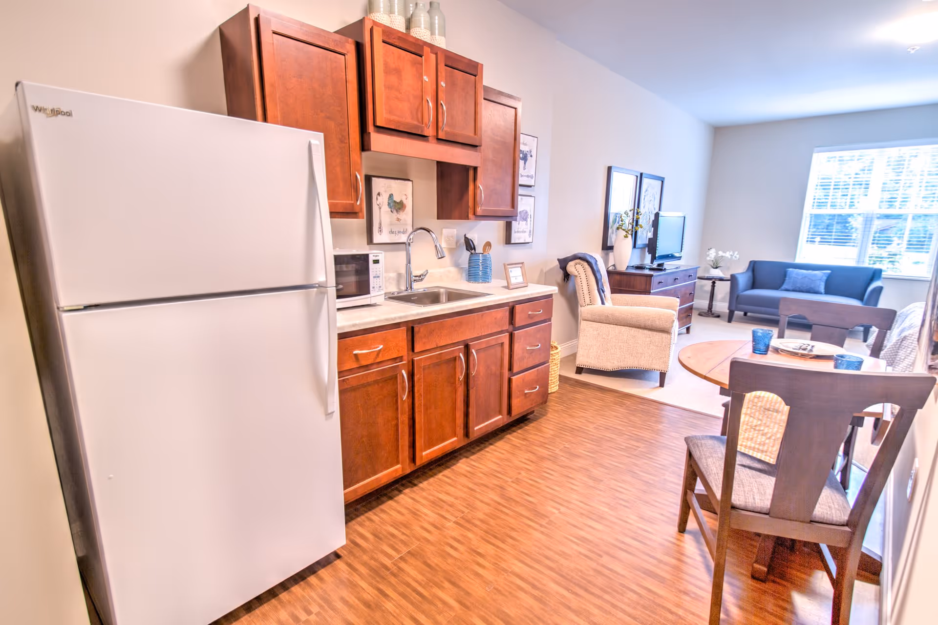 A bright and clean senior living apartment interior featuring a white Whirlpool refrigerator, wooden cabinets, a microwave, and a sink in the kitchen area. Adjacent to the kitchen is a small dining table with chairs. In the background, there is a living area with a beige armchair, a blue loveseat, a wooden TV stand with a television, and a large window letting in natural light.