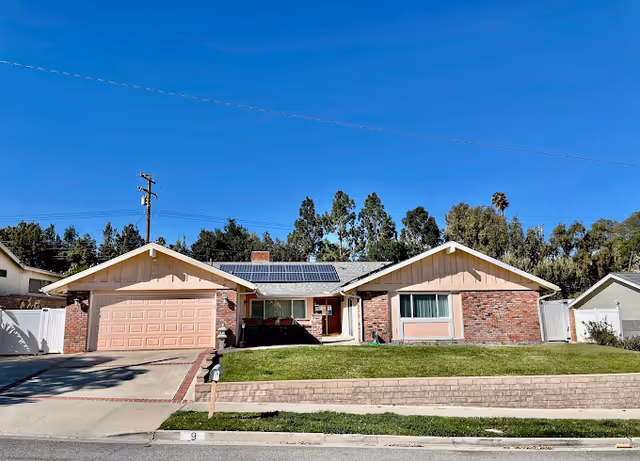 Single-story ranch-style house with a two-car garage, front lawn, and solar panels on the roof under a clear blue sky.