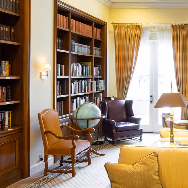 A cozy library or sitting area with wooden bookshelves filled with books, a globe on a stand, a brown leather armchair, a purple leather armchair, yellow curtains framing glass doors, and a yellow sofa with a matching pillow in the foreground.