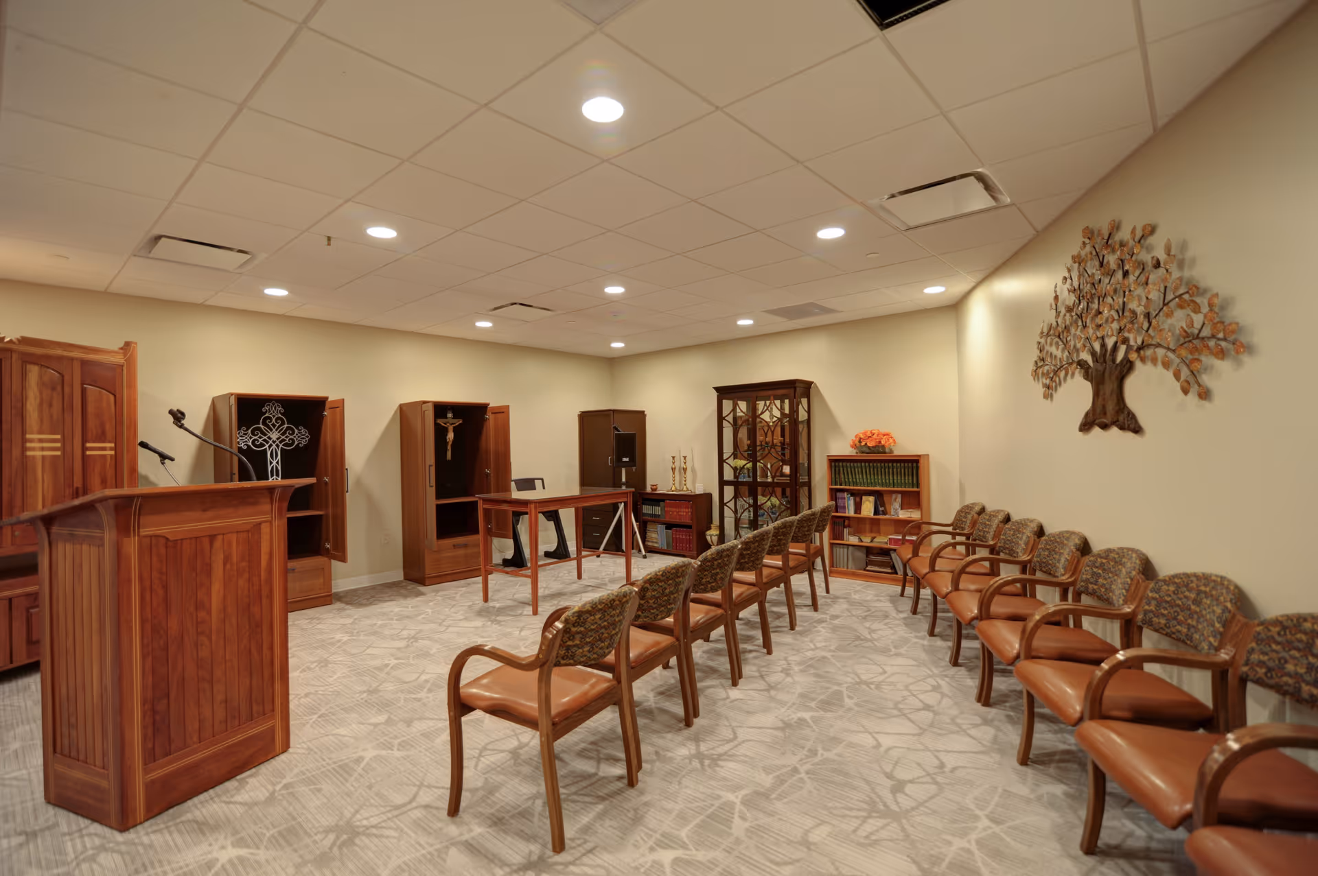 A small chapel or meditation room with rows of wooden chairs with cushioned seats arranged facing a wooden podium and a table. The room has beige walls, a patterned carpet, and a decorative metal tree wall art. There are wooden cabinets with religious symbols and a bookshelf with books and flowers.