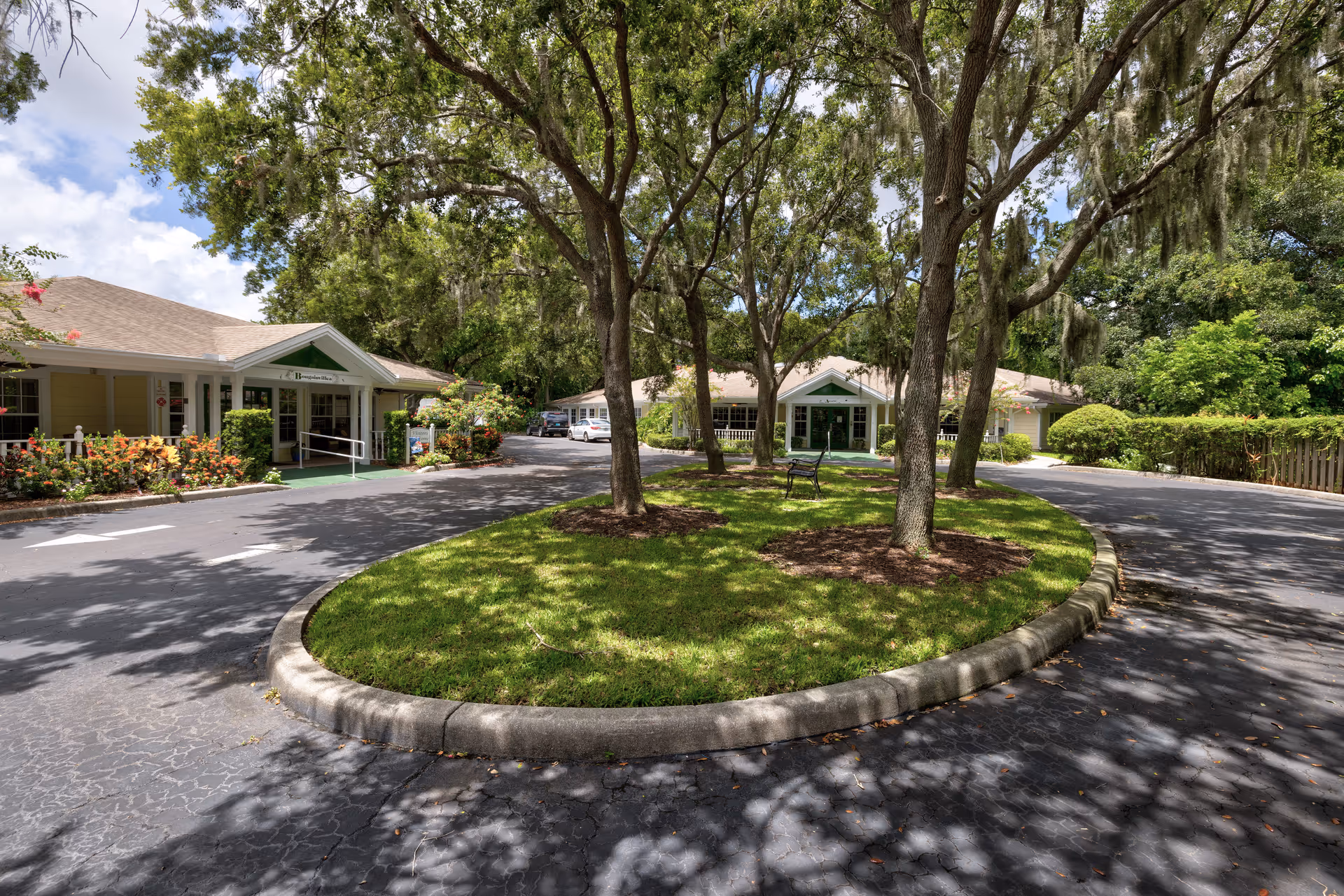 View of the entrance area of Discovery Commons At Bradenton, showing a circular driveway with a grassy island in the center featuring several trees and a bench. The building has a light-colored exterior with green accents and is surrounded by lush greenery and flowers under a partly cloudy sky.