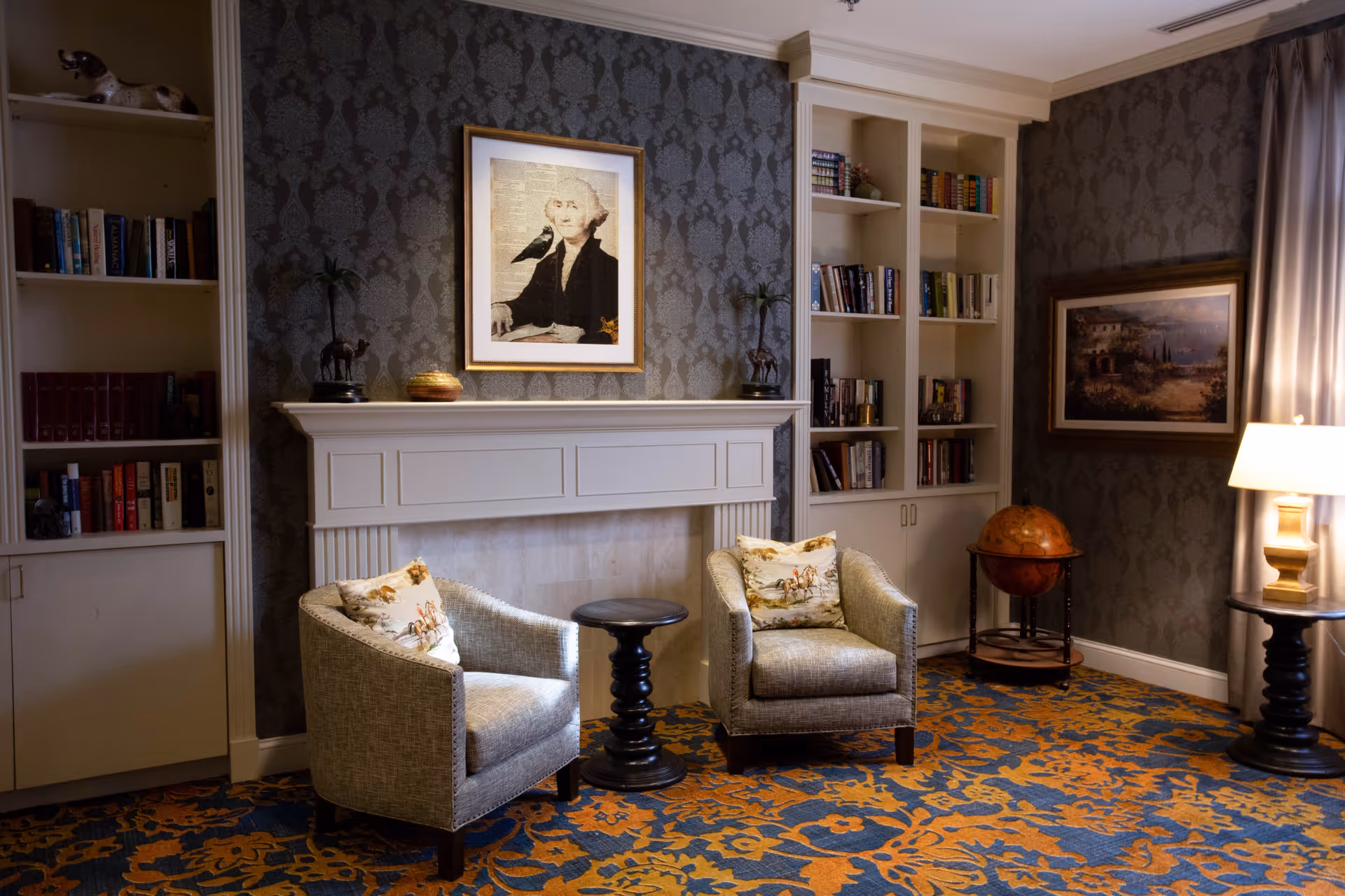 A cozy living room area with two upholstered armchairs featuring decorative pillows, a small round black side table between them, and a white fireplace with a framed portrait above it. Built-in bookshelves flank the fireplace, filled with books. A globe on a stand and a table lamp on a round side table are visible on the right side. The room has patterned wallpaper and a blue and orange floral carpet.