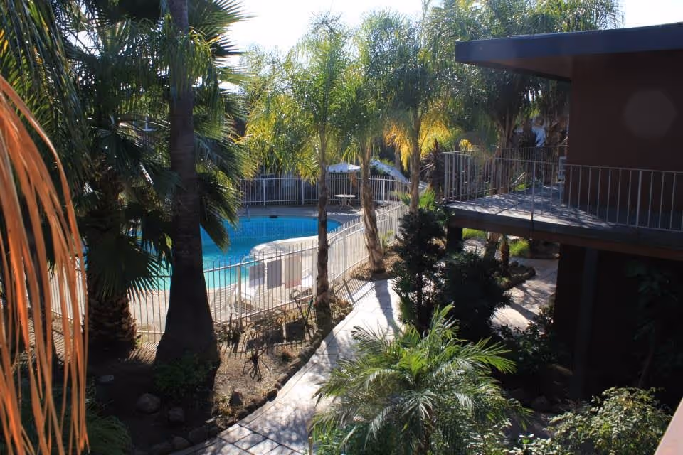 Courtyard with palm trees, a fenced swimming pool, and a walkway beside a two-story building with balconies.