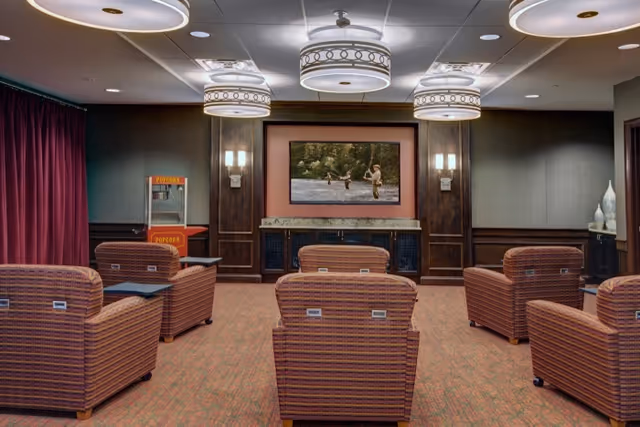 A cozy media room with several striped upholstered armchairs arranged in rows facing a large flat-screen TV mounted on a wood-paneled wall. The room features warm lighting with three decorative ceiling lights, wall sconces, a popcorn machine in the corner, and a carpeted floor. Dark curtains cover the windows on the left side.