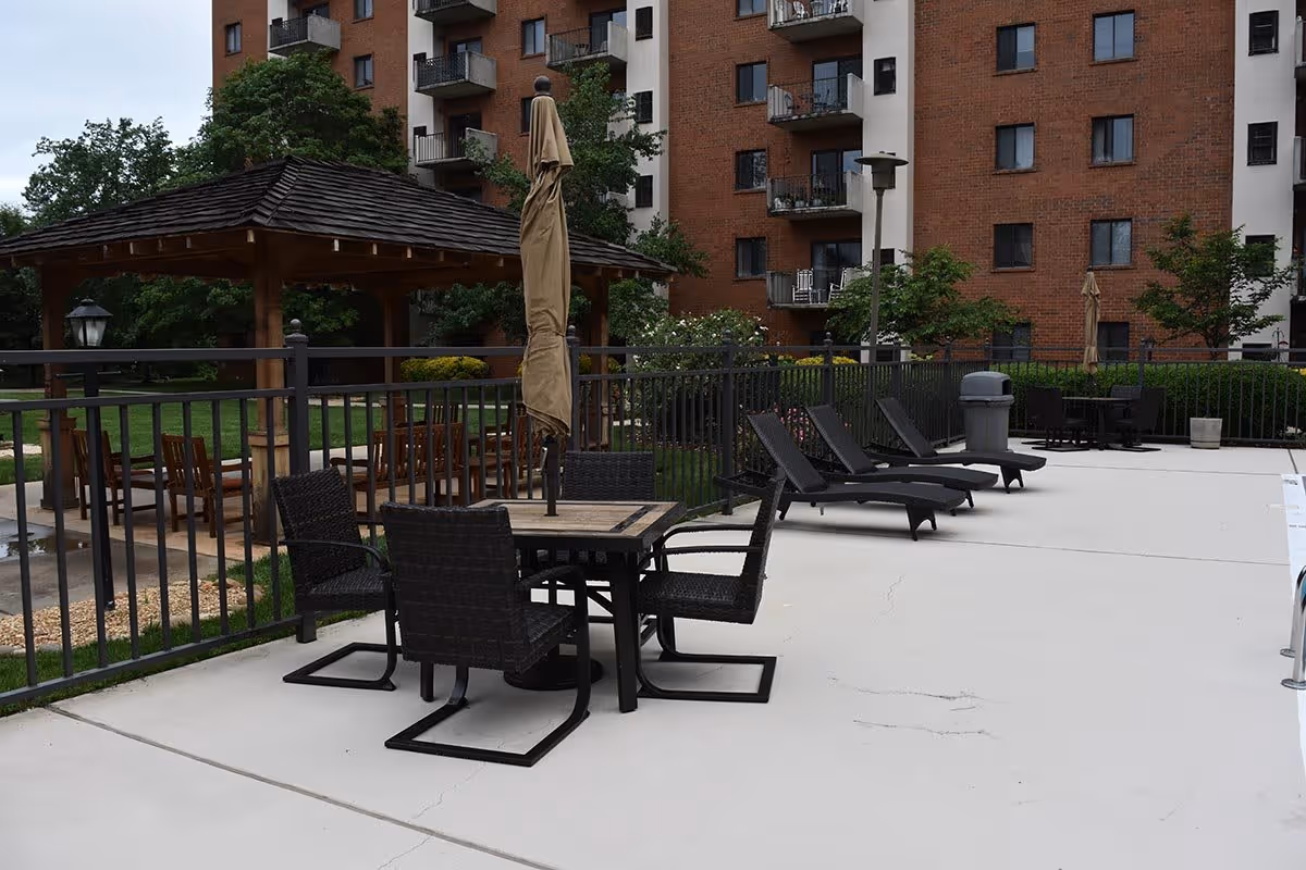 Outdoor patio with tables, lounge chairs, umbrellas and a wooden gazebo in front of a multi-story brick apartment building.