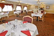 Formal dining room with round tables set with white linens and red napkins, chairs, patterned carpet, and floral wallpaper.