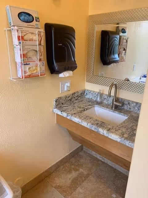 Small bathroom sink area with a granite countertop, wall-mounted paper towel dispenser, mirror, and glove dispenser on beige walls.