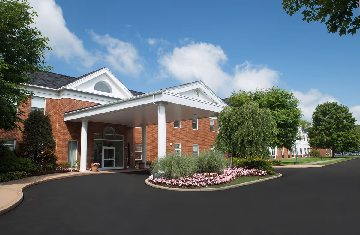 Front exterior view of a red brick senior living facility with a white portico entrance, surrounded by landscaped greenery and pink flowers under a blue sky with some clouds.