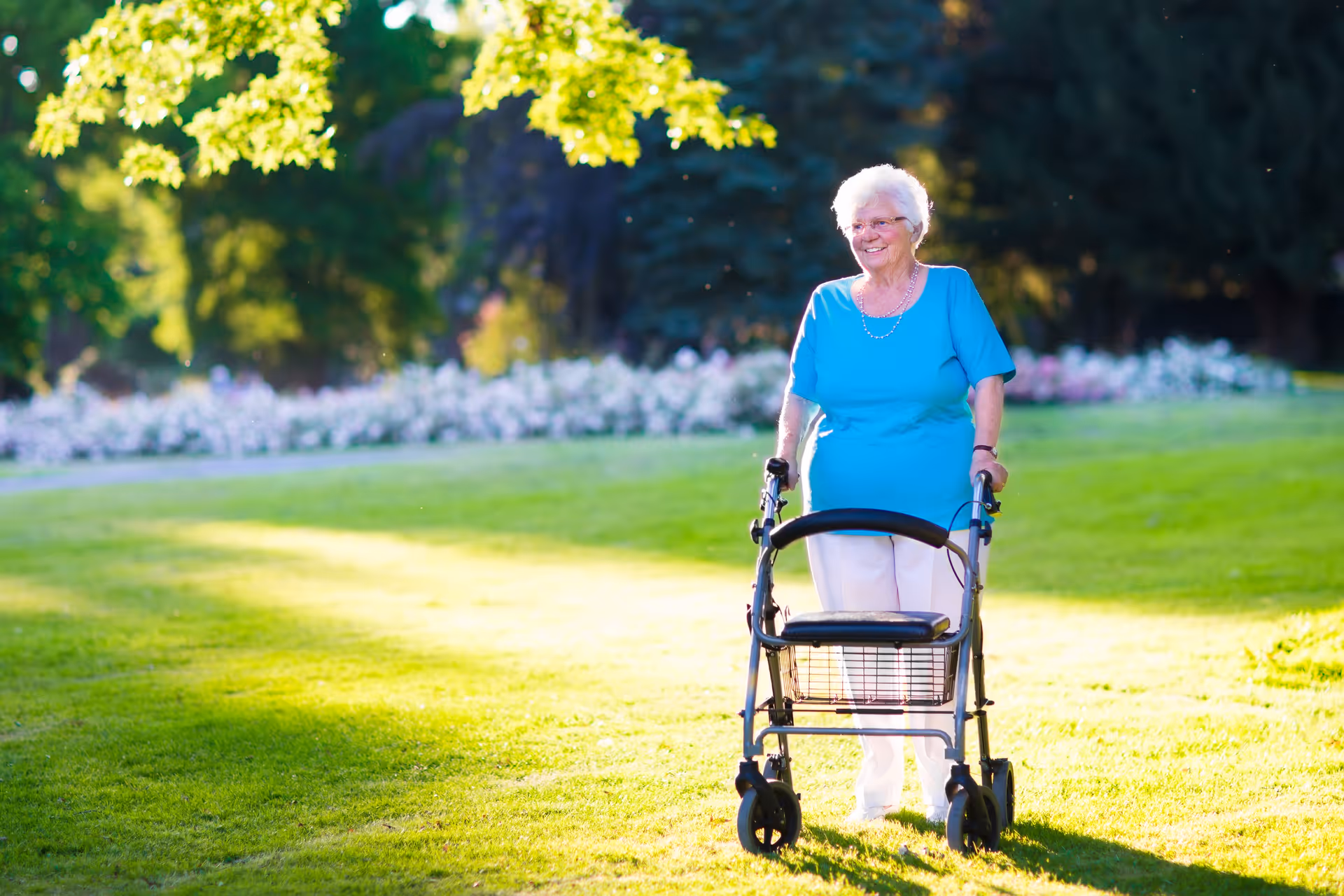 An elderly woman with white hair wearing a blue shirt and white pants is standing outdoors on a grassy area using a walker. She is smiling and there are trees and flowers in the background with sunlight filtering through the leaves.