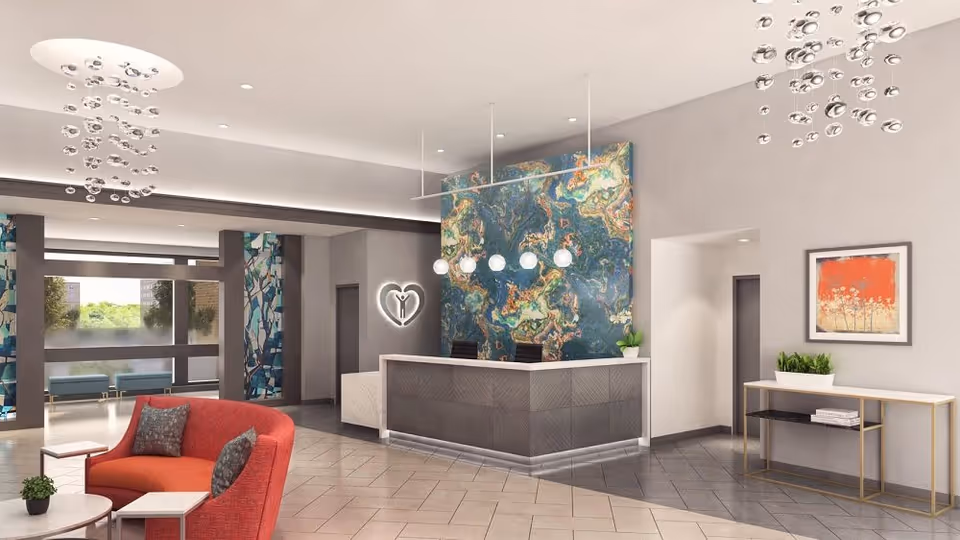 A modern reception area with a front desk featuring a colorful abstract wall behind it, two black chairs, and hanging globe pendant lights. To the left, there is a red curved sofa with two pillows and a small round table with a plant. The floor is tiled, and there are large windows with blue patterned curtains letting in natural light. On the right wall, there is a framed artwork and a console table with plants and books. The ceiling has recessed lighting and decorative hanging light fixtures.