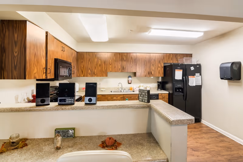 Communal kitchen with wood cabinets, a black refrigerator, sink and countertop with small appliances and speakers.