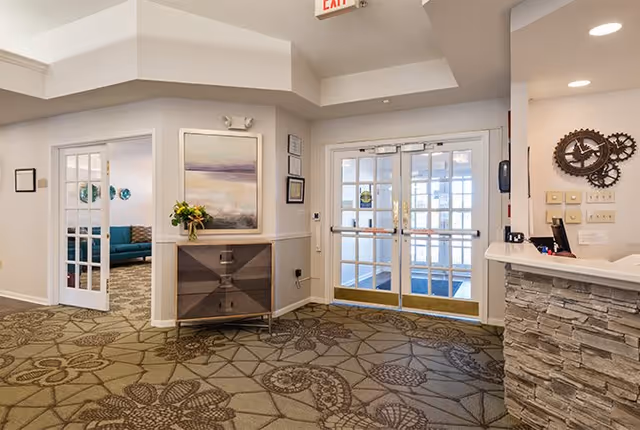 Interior view of a senior living facility entrance area with double glass doors, a patterned carpet, a small cabinet with a vase of flowers, and a reception desk with stone facade. A room with a teal sofa is visible through an open door on the left.