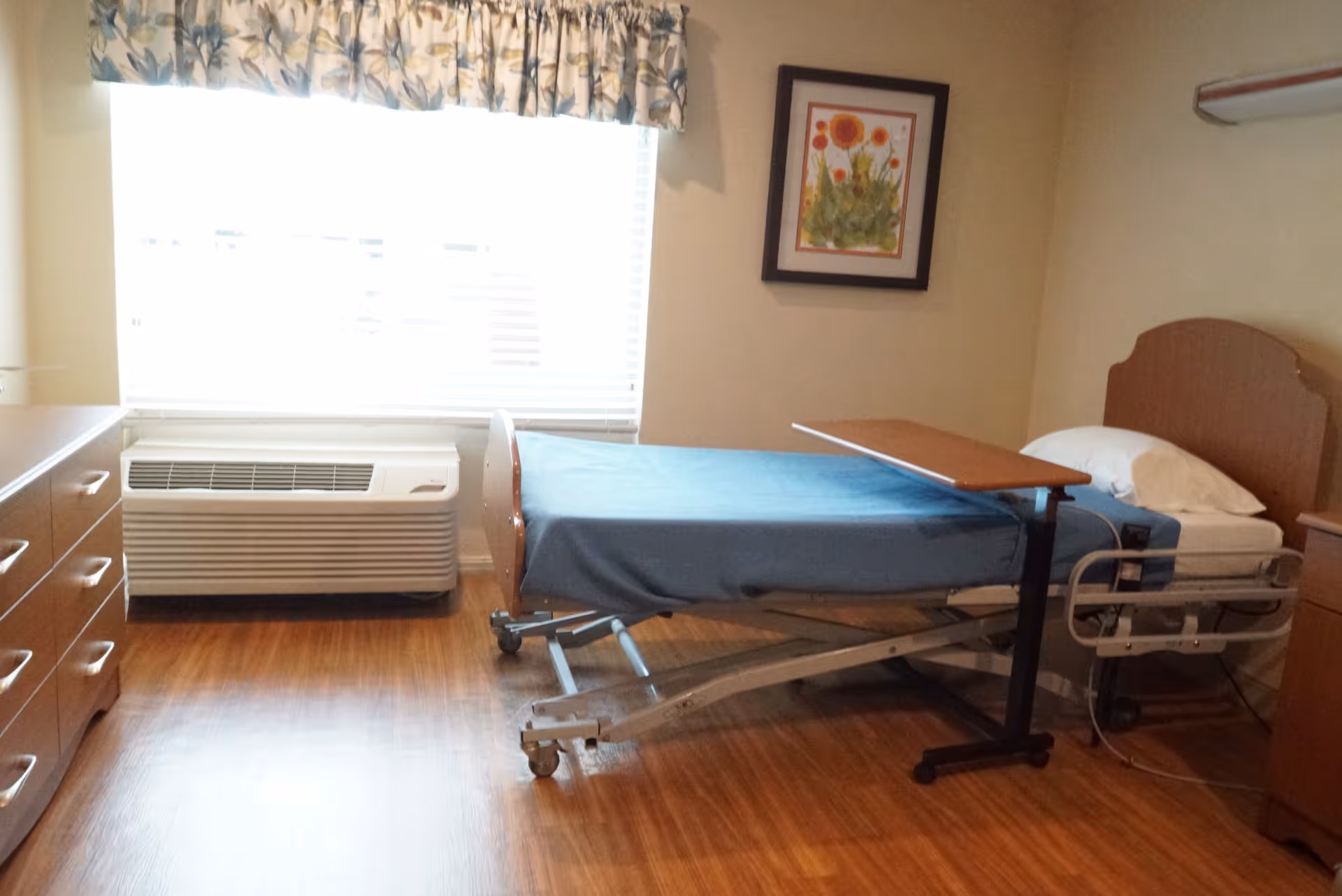 A simple bedroom in a senior living facility featuring a hospital-style bed with a blue blanket, a wooden overbed table, a wooden dresser, a framed floral painting on the wall, a window with blinds and floral valance, and an air conditioning unit below the window.