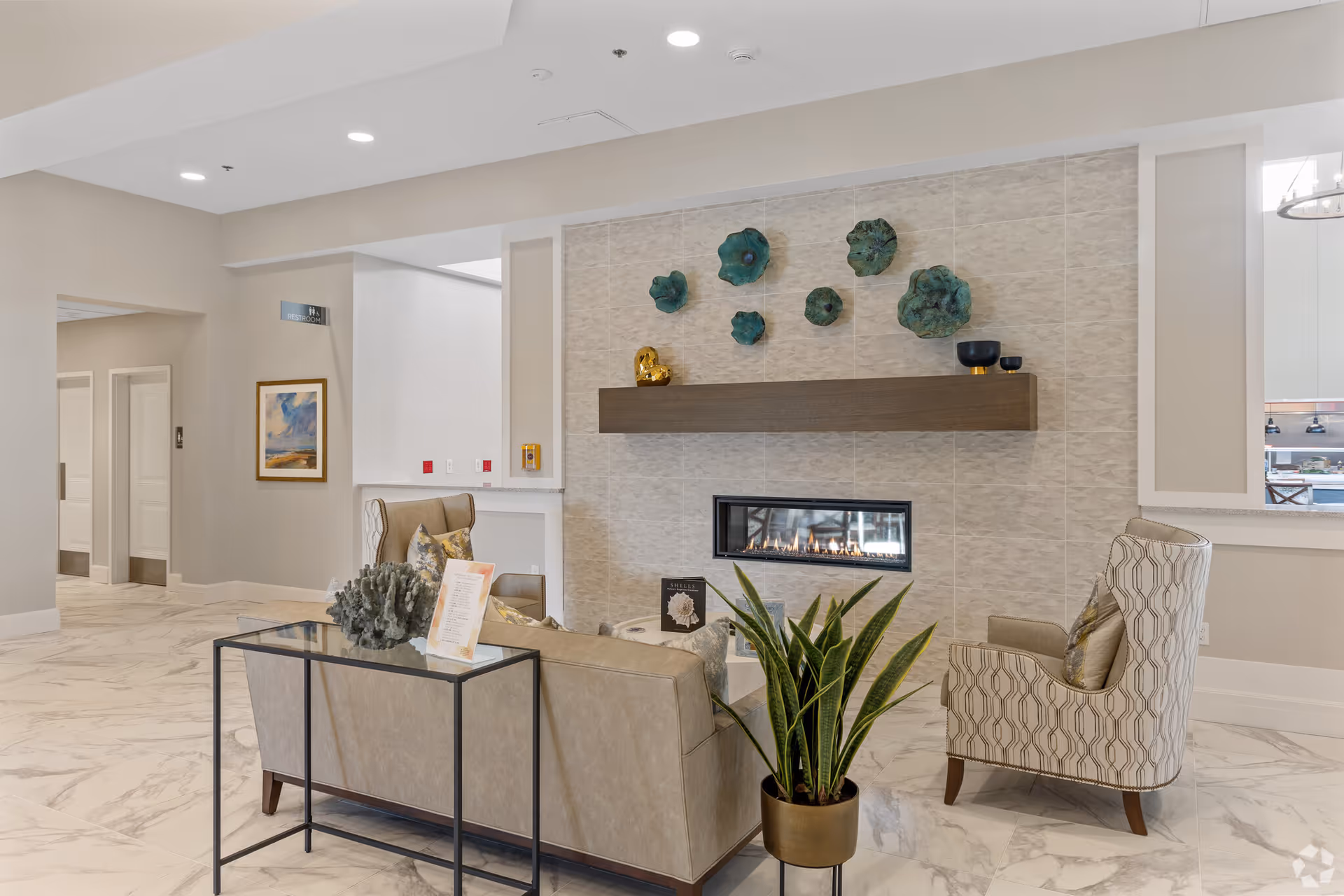A modern living room area with a beige sofa and patterned armchair facing a wall-mounted linear gas fireplace. Above the fireplace is a wooden mantel with decorative items and green wall art. The floor is covered with white marble tiles, and there is a potted plant near the sofa. The room has recessed lighting and neutral-colored walls.