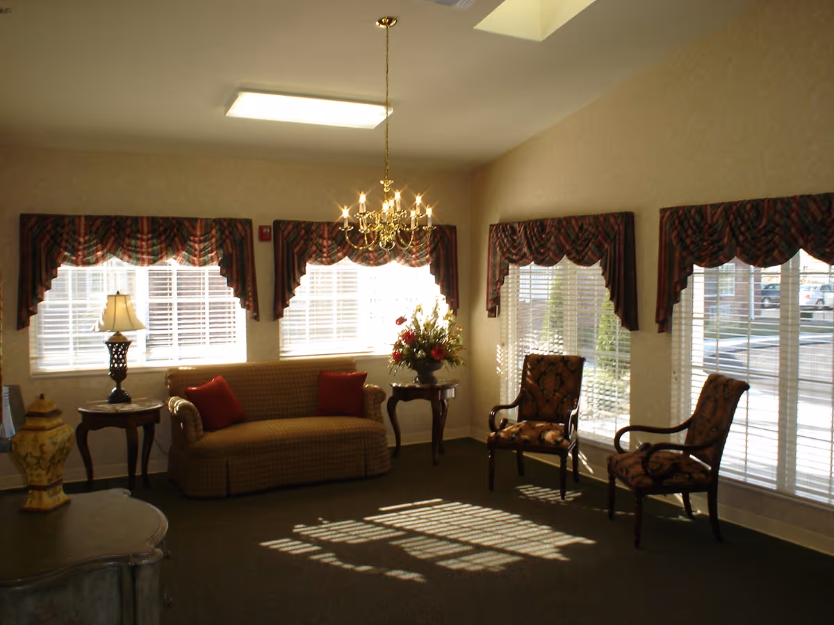 A cozy living room area with a beige couch adorned with two red pillows, two patterned armchairs, a side table with a lamp, and a small table with a floral arrangement. The room has large windows with blinds and plaid valances, a chandelier hanging from the ceiling, and sunlight casting shadows on the carpeted floor.
