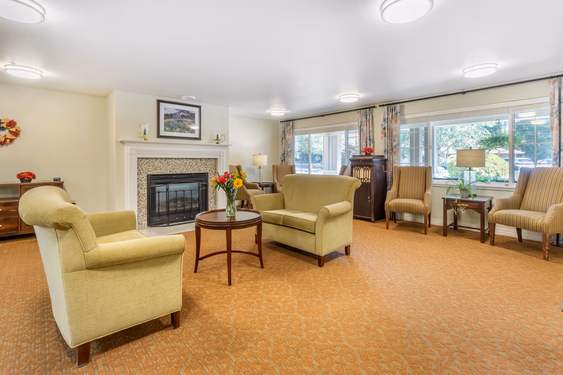 Sunny common living room with green upholstered chairs and a loveseat around a coffee table in front of a fireplace and large windows.