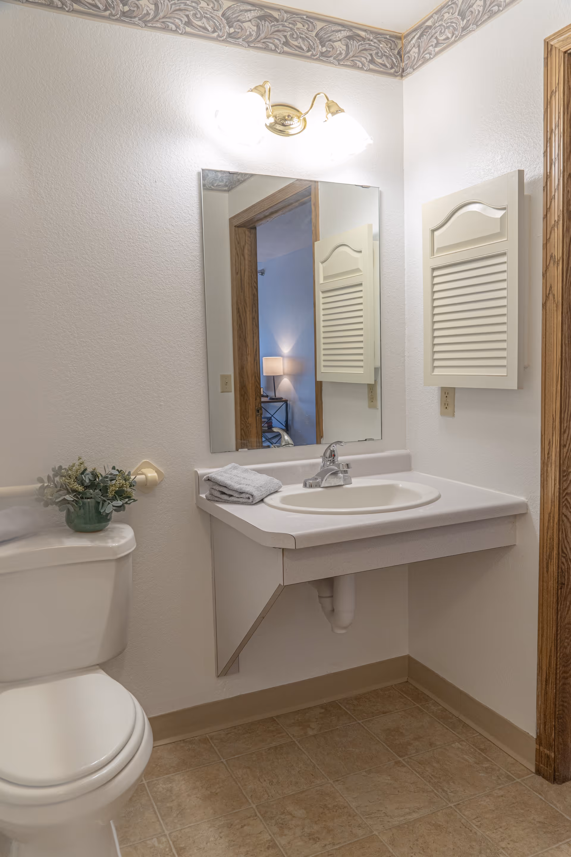 A clean bathroom with a white toilet and a wall-mounted sink with a faucet. Above the sink is a large mirror reflecting part of an adjacent room with a lamp. There is a decorative border near the ceiling and a small green plant on the toilet tank. The floor is tiled in a light brown color.