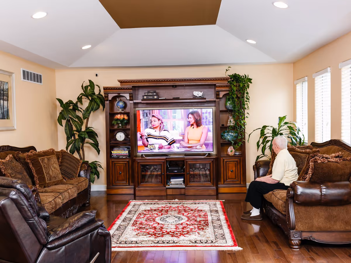 A cozy living room with wooden flooring, a patterned red and white rug, and brown upholstered sofas. An elderly person is sitting on one of the sofas watching a television mounted in a wooden entertainment center. The room has several green plants and windows with white blinds letting in natural light.