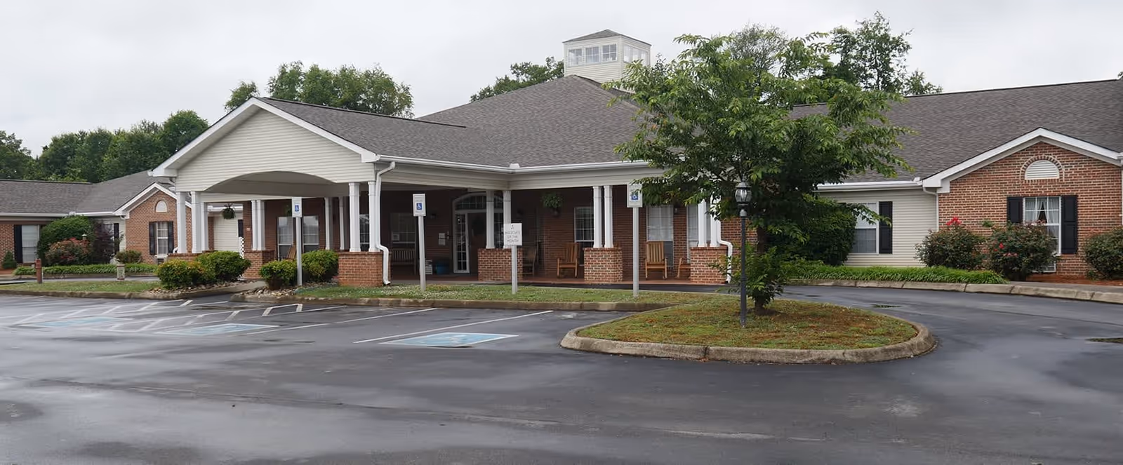 Exterior view of a single-story senior living facility building with a covered entrance, brick and siding facade, surrounded by a parking lot with marked handicap parking spaces and a small landscaped island with a tree in front.