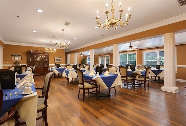A spacious dining room with wooden floors, multiple tables covered with white and blue tablecloths, each set with napkins, cups, and silverware. The room features elegant chandeliers, large windows letting in natural light, white columns, and a wooden cabinet against the far wall.