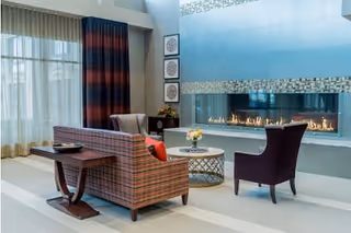 A cozy senior living community lounge area with a modern linear fireplace set in a blue tiled wall. The room features a plaid upholstered sofa with a wooden side table, a round coffee table with a flower arrangement, and a purple armchair. Large windows with sheer and dark curtains allow natural light to fill the space.