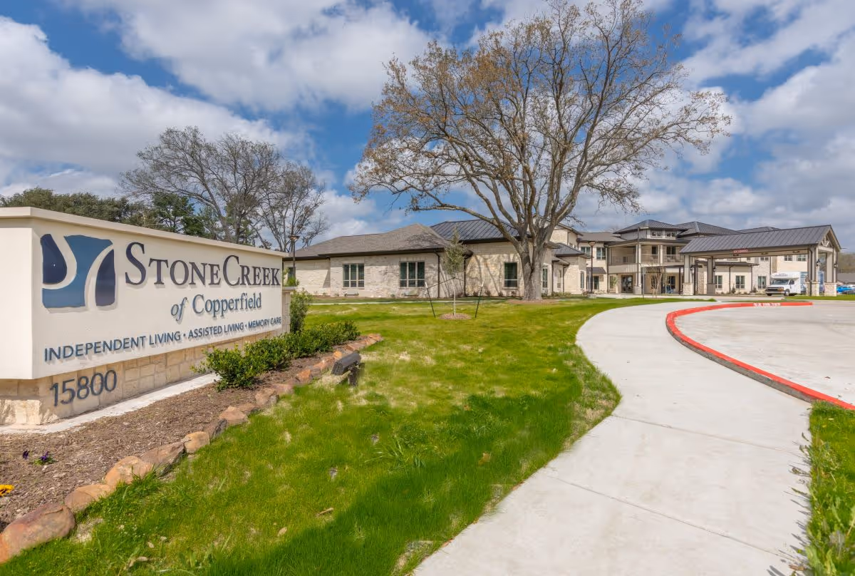Exterior view of StoneCreek of Copperfield Senior Living facility with a large sign displaying the name and services including Independent Living, Assisted Living, and Memory Care. The building is surrounded by green grass, trees, and a curved driveway under a partly cloudy sky.