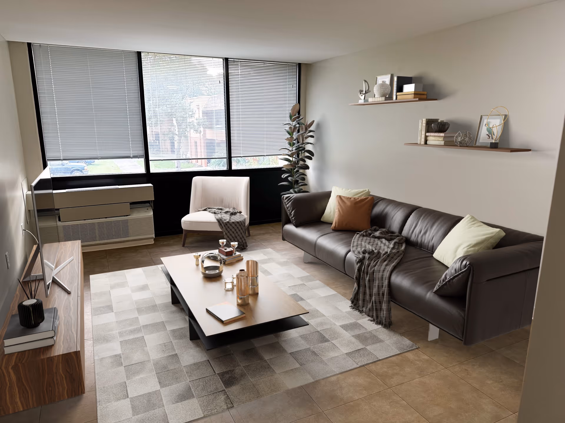 A modern living room with a large window covered by blinds, a dark brown leather sofa with pillows and a throw blanket, a light-colored armchair with a throw, a wooden coffee table with decorative items, a patterned rug, two floating shelves with books and decor on the wall, and a TV on a wooden stand.