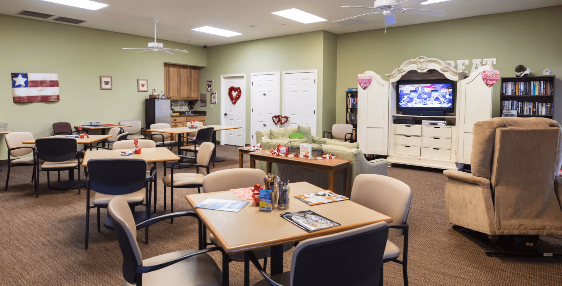 A bright communal activity room with multiple tables and chairs, a TV cabinet, sofas and a small kitchenette in the back.