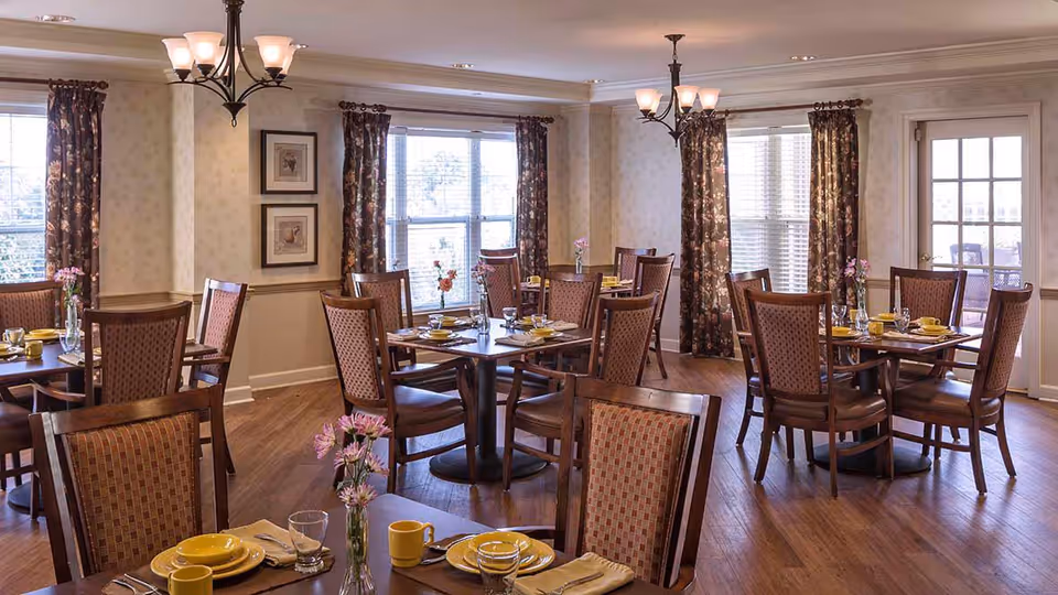 A dining room with multiple wooden tables and chairs arranged neatly. Each table is set with yellow plates, cups, and silverware, along with small vases containing pink flowers. The room has large windows with floral curtains, wooden flooring, and soft lighting from ceiling fixtures.