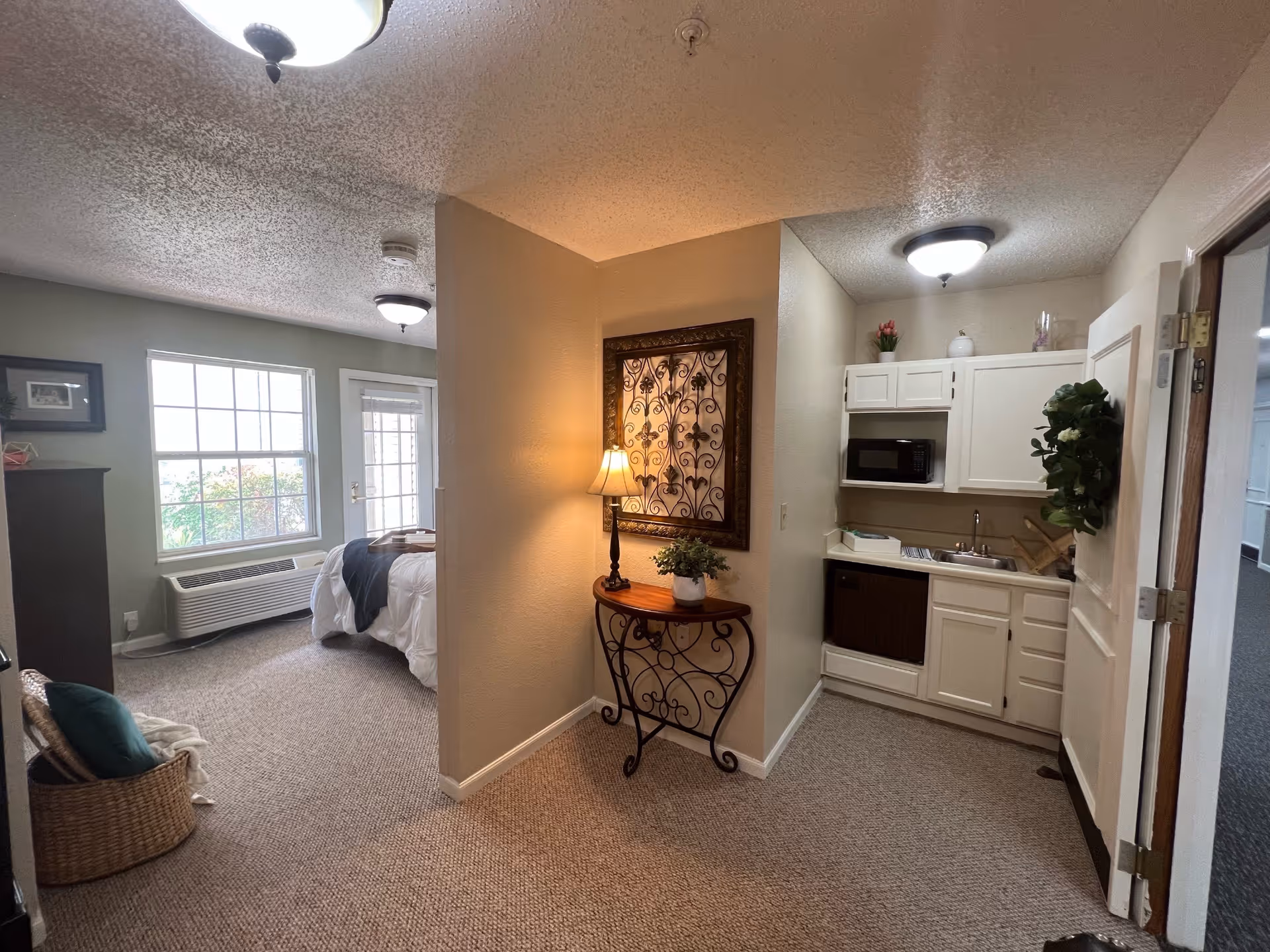 Interior view of a senior living facility apartment showing a small kitchenette with white cabinets, a microwave, and a sink on the right. In the center, there is a decorative table with a lamp and a framed wall art above it. On the left, a bedroom area is visible with a bed, a window, and a door leading outside. The room is carpeted and well-lit with ceiling lights.