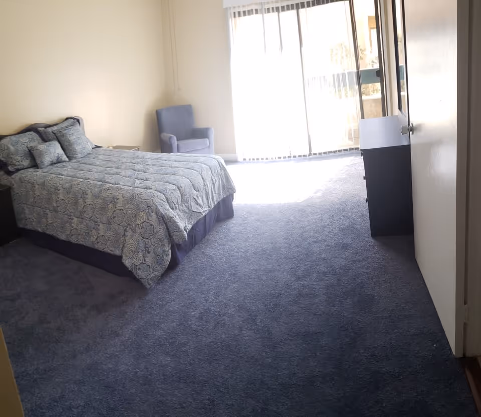 A bedroom with a bed covered in a blue patterned comforter and matching pillows, a blue armchair in the corner, a dresser near the door, and a large sliding glass door letting in natural light.