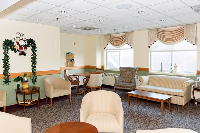 A bright senior living facility common area with beige and patterned armchairs, a striped sofa, wooden coffee tables, and a small round dining table with chairs near the windows. The walls are painted light yellow with a green lower half and wooden trim. There is a decorative Santa Claus hanging on a green leafy arch on one wall, and the windows have beige valances.