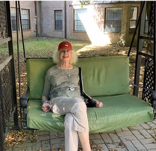 An elderly woman wearing a red cap, gray long-sleeve shirt, and light-colored pants is sitting on a green cushioned outdoor swing. The swing is on a paved area with a brick building and windows in the background, along with some grass and fallen leaves on the ground.