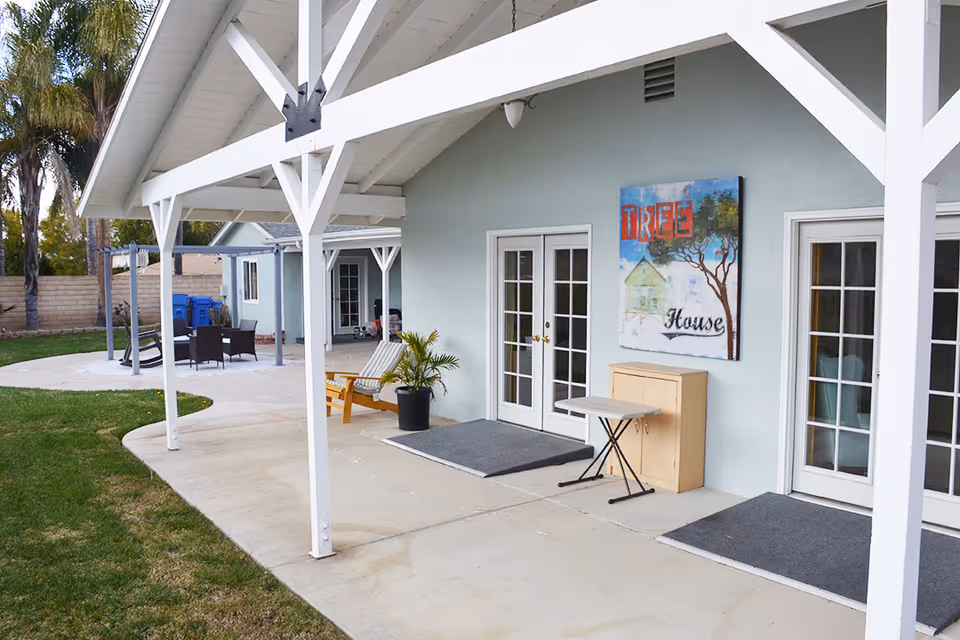 Covered patio with white beams, French doors, outdoor seating, and lawn beside a light blue building.