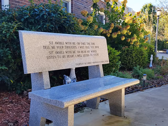 A stone bench with an engraved poem is placed outdoors near a brick building and surrounded by green bushes and plants. The bench inscription reads: 'Sit awhile with me, oh take the time / Tell me your thoughts, I will tell you mine / Sit awhile with me, oh hear my words / Listen to my heart, I will listen to yours.'