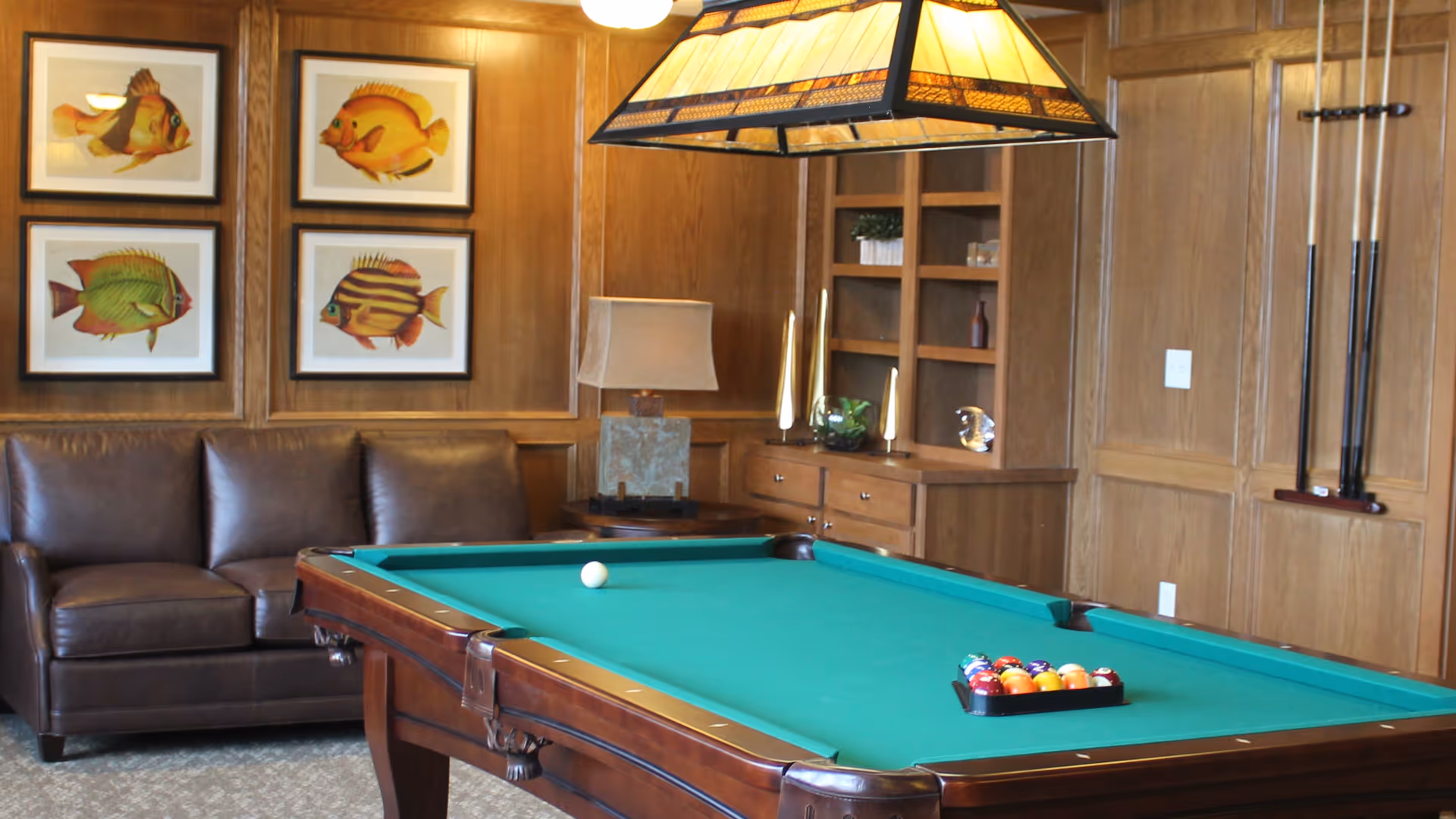 A cozy game room featuring a green felt pool table with balls racked and a cue ball placed on the table. Behind the pool table is a brown leather couch, wooden paneled walls with four framed fish illustrations, a table lamp on a side table, built-in wooden shelves with decorative items, and pool cues mounted on the wall.