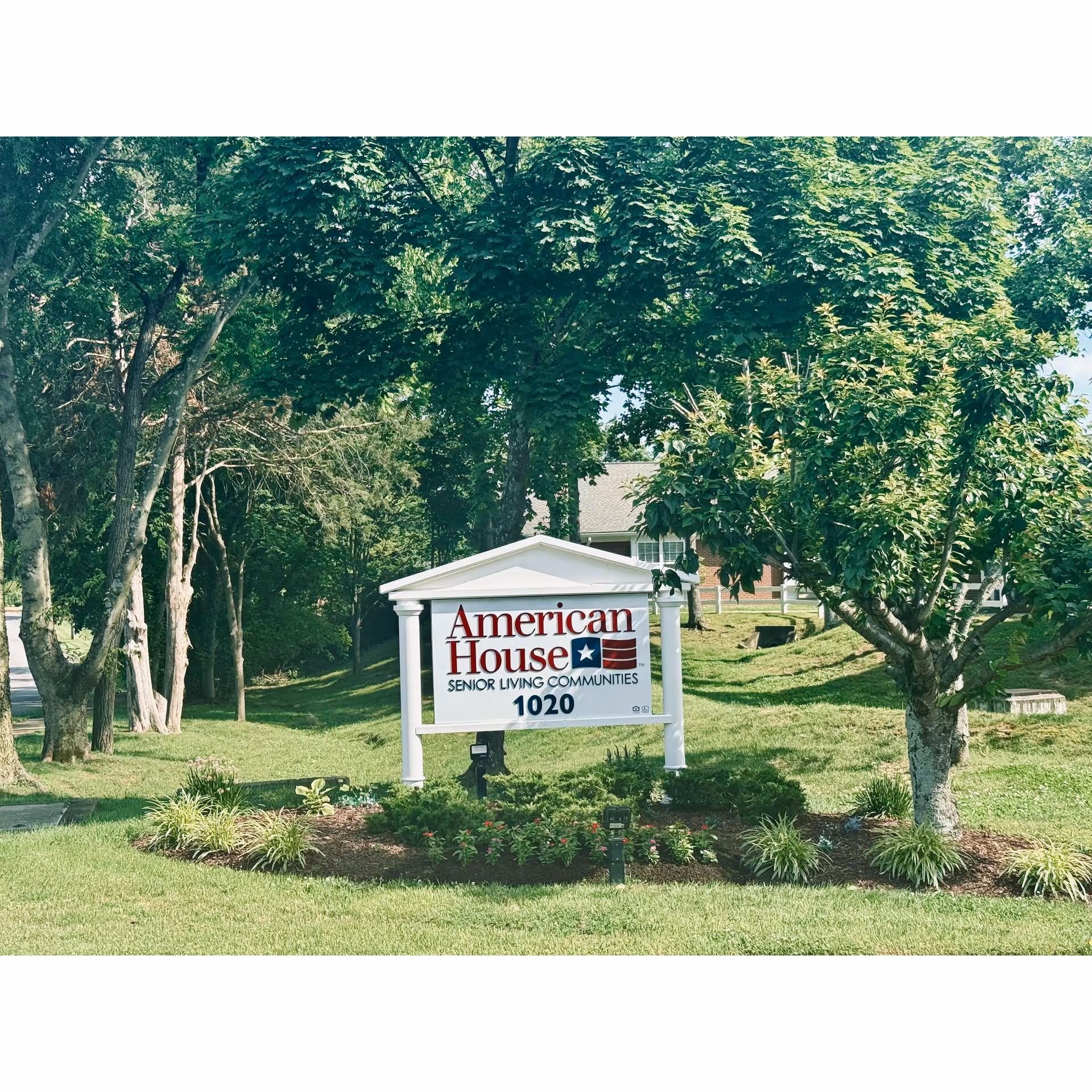 Outdoor sign reading "American House Senior Living Communities" on a manicured lawn with trees and a building in the background.