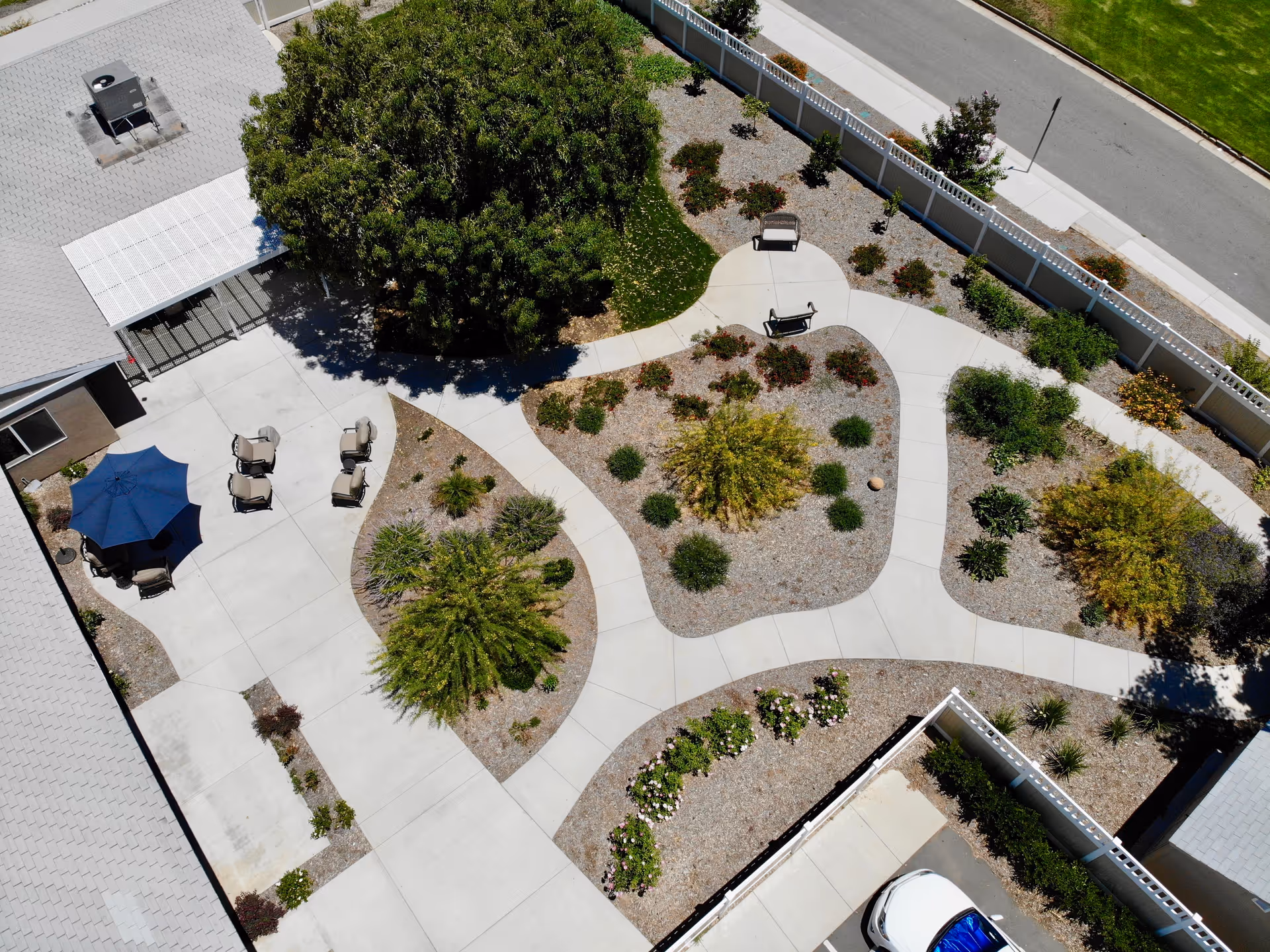 Aerial view of an outdoor garden area at The Hills of Stillman featuring concrete walkways, landscaped planting beds with shrubs and small trees, several benches, and a patio area with chairs and a blue umbrella. The area is enclosed by a white fence and adjacent to a parking lot and building.