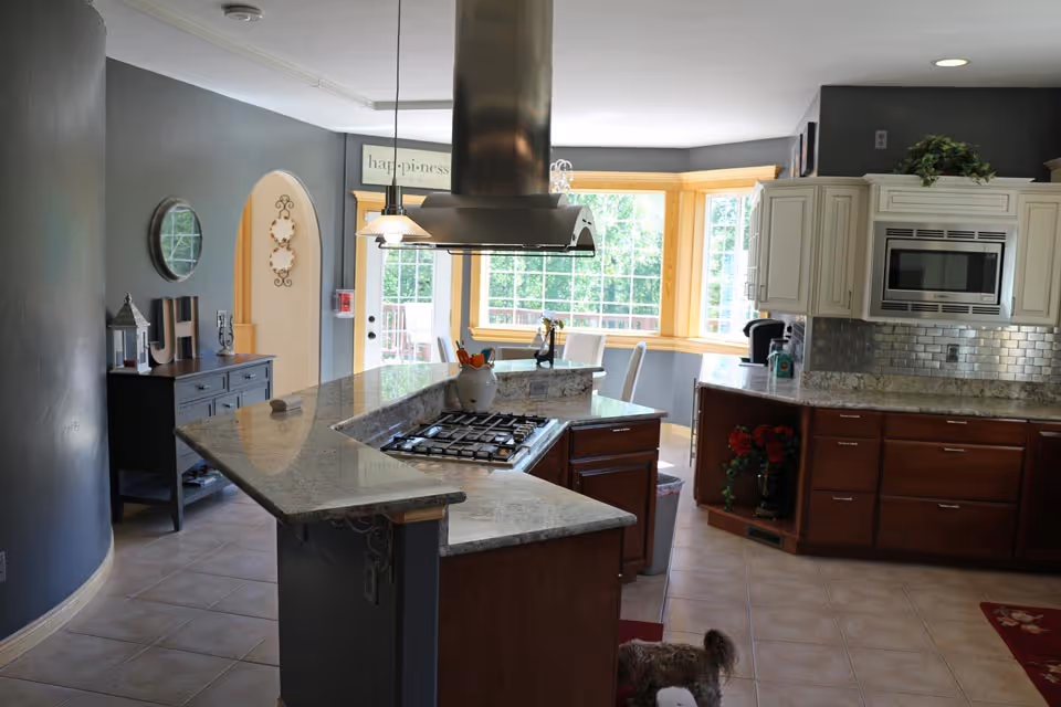 A modern kitchen with a large granite countertop island featuring a built-in gas stove and a stainless steel range hood above it. The kitchen has white upper cabinets and dark wood lower cabinets, a microwave built into the cabinetry, and a tiled backsplash. There is a small dog standing on the tiled floor near the island. In the background, there is a dining area with large windows letting in natural light and a sign above the door that reads 'happiness'.