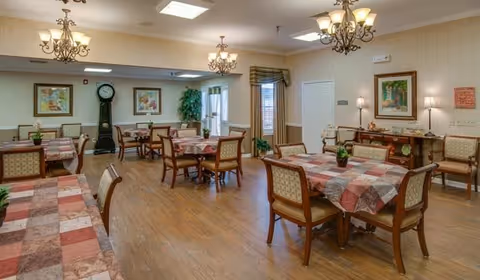 A spacious dining room with several wooden tables covered with checkered tablecloths and surrounded by cushioned chairs. The room features hardwood flooring, three ornate chandeliers, framed artwork on the walls, a grandfather clock, and a sideboard with decorative items. There are windows with curtains and a door leading to another area.