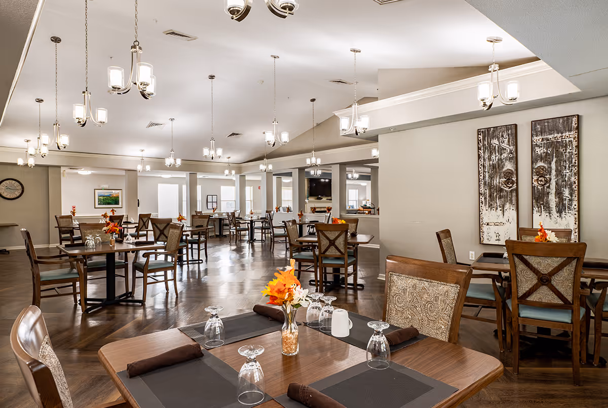 Spacious dining room with multiple wooden tables and chairs arranged neatly. Each table is set with upside-down wine glasses, brown napkins, and small floral centerpieces with autumn-colored flowers. The room features a high ceiling with multiple hanging light fixtures, wooden flooring, and neutral-colored walls adorned with rustic artwork and a clock.