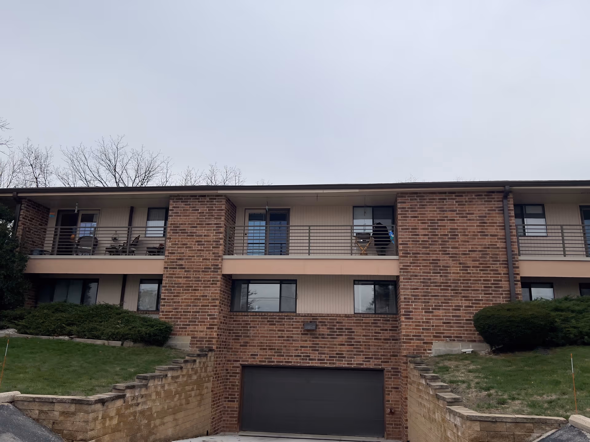 Exterior view of a two-story brick building with a garage door at the center bottom. The upper floor has balconies with metal railings and patio furniture. There are bushes and grass on either side of the building, and leafless trees are visible in the background under a cloudy sky.