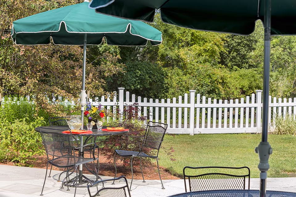 Outdoor patio area with black metal table and chairs under green umbrellas. The table has a pitcher of lemonade, two glasses, a vase with colorful flowers, and red placemats. A white picket fence and green trees and bushes are visible in the background.