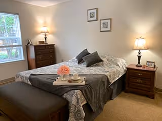 A cozy bedroom with a neatly made bed featuring a patterned bedspread and several pillows. There is a wooden nightstand with a lamp and a framed photo on the right side of the bed, and a wooden dresser with a lamp and decorative items near a window on the left. Two framed pictures hang on the wall above the bed. A cushioned bench is placed at the foot of the bed.
