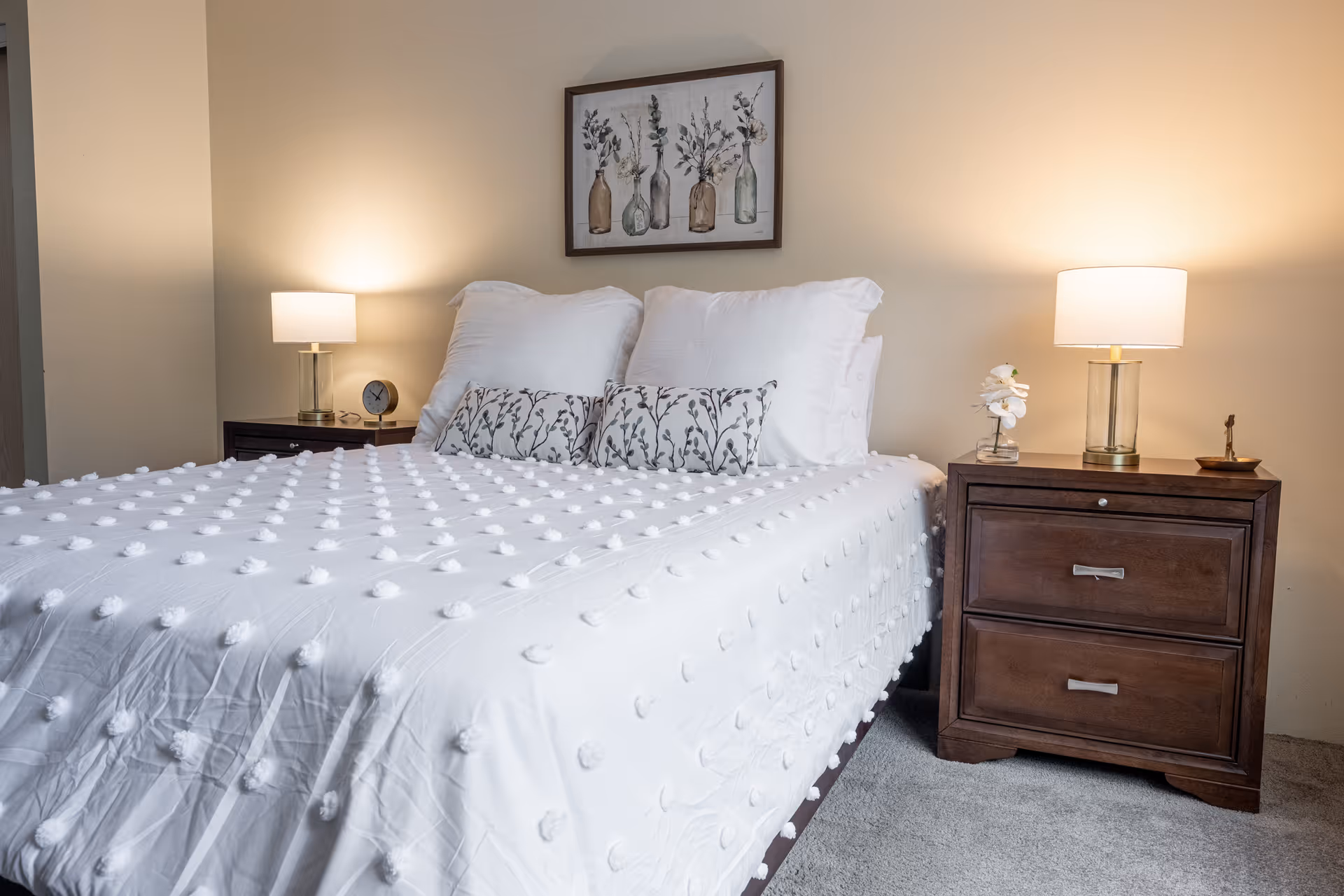 A neatly made bed with a white textured comforter and two decorative pillows with a botanical pattern. On either side of the bed are dark wooden nightstands, each with a lamp. One nightstand also has a small clock, and the other has a small decorative item. Above the bed hangs a framed artwork featuring bottles with plants inside. The room has beige walls and carpeted flooring.