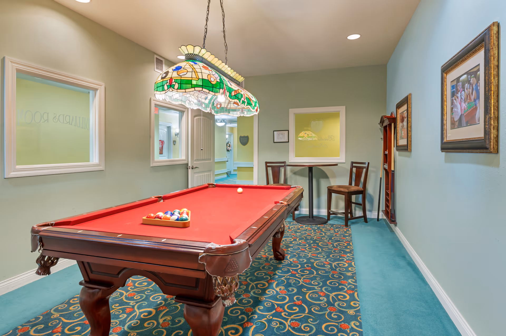 Interior view of a game room with a red felt pool table, a colorful stained glass hanging light above it, a small round table with two chairs against the wall, framed pictures on the blue wall, and windows looking into adjacent rooms.