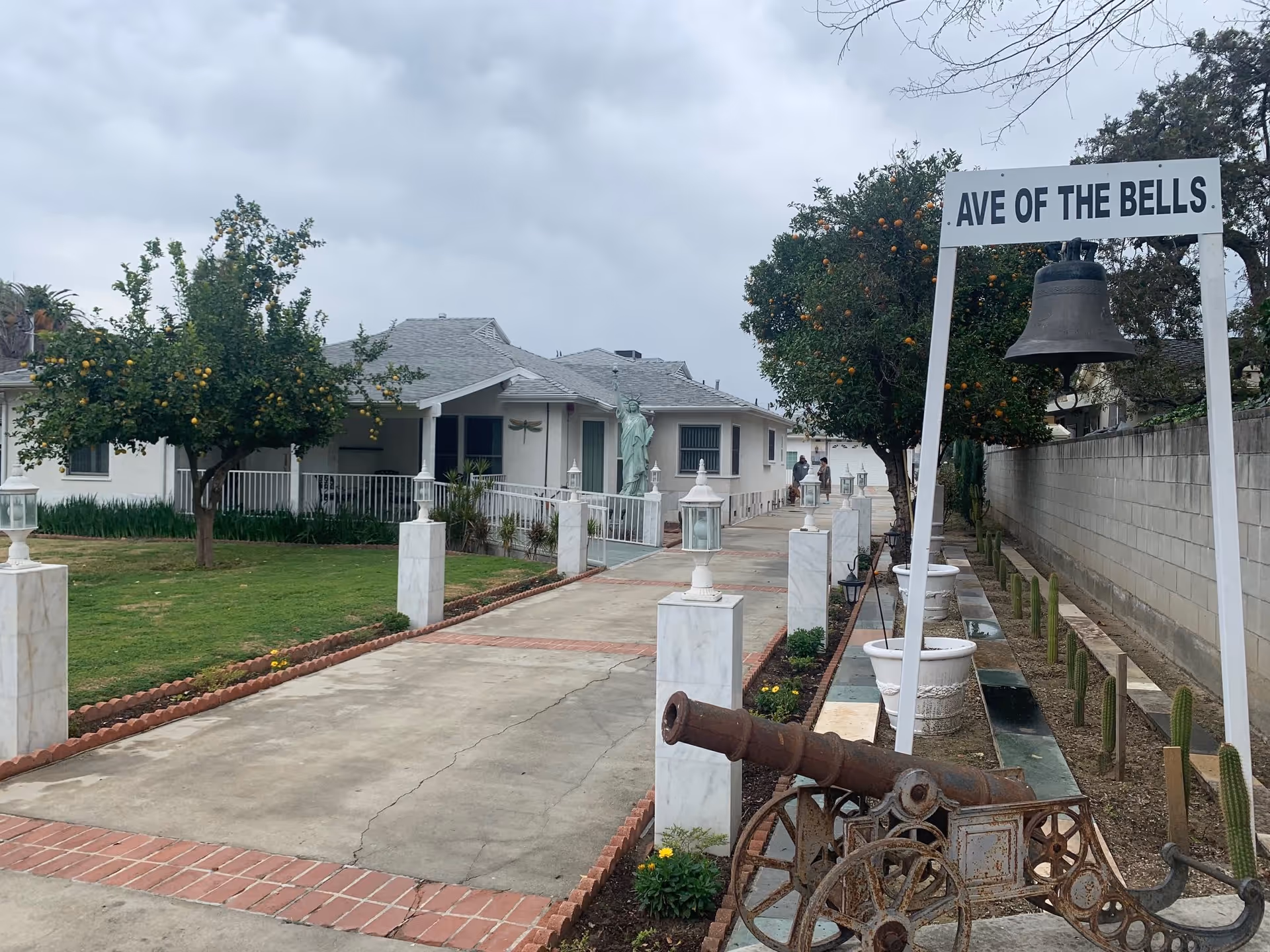 Outdoor view of a senior living facility named Ave of the Bells featuring a paved walkway lined with white lamp posts and orange trees. A large bell hangs from a white frame with the facility name above it, and an old cannon is positioned nearby. The building is a single-story structure with a gray roof and a small statue resembling the Statue of Liberty near the entrance. Two people are seen walking in the background.