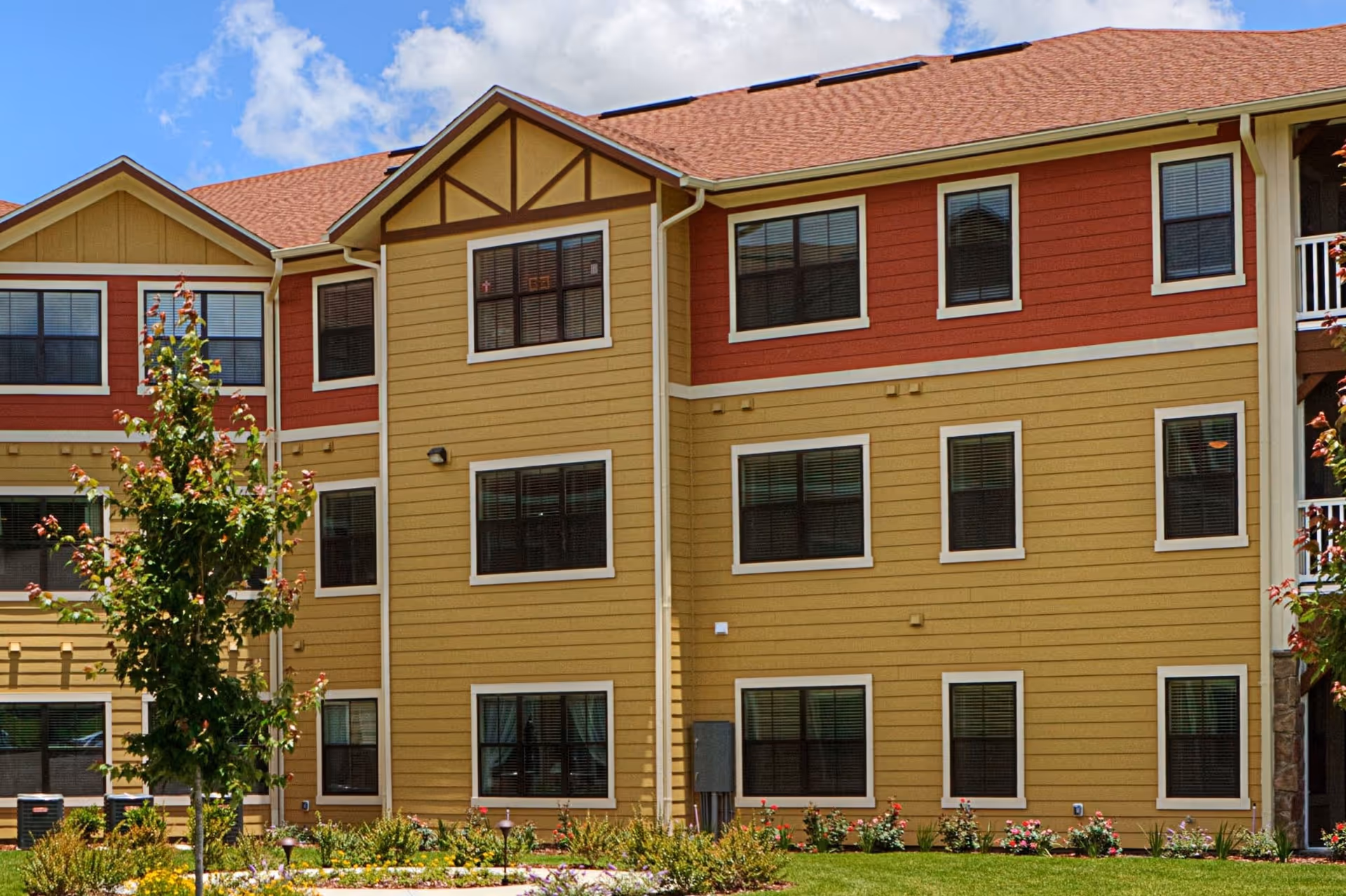 Exterior view of a multi-story senior living facility building with red and yellow siding, multiple windows, a landscaped garden with flowers and small trees, and a partly cloudy blue sky.